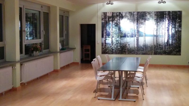 Kuva tyhjästä salista jonka keskellä seisoo kahdeksan hengen pöytä. Taustalla seinän kokoinen valokuva suomalaisesta rantakoivikosta.