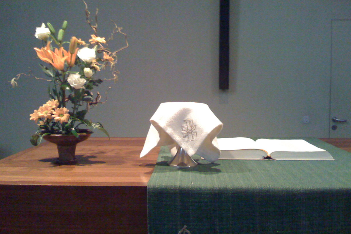 Lukas-kirkon alttari. Etualalla liinalla peittty ehtoollismalja. Taustalla avattu raamattu ja kukka-asetelma.